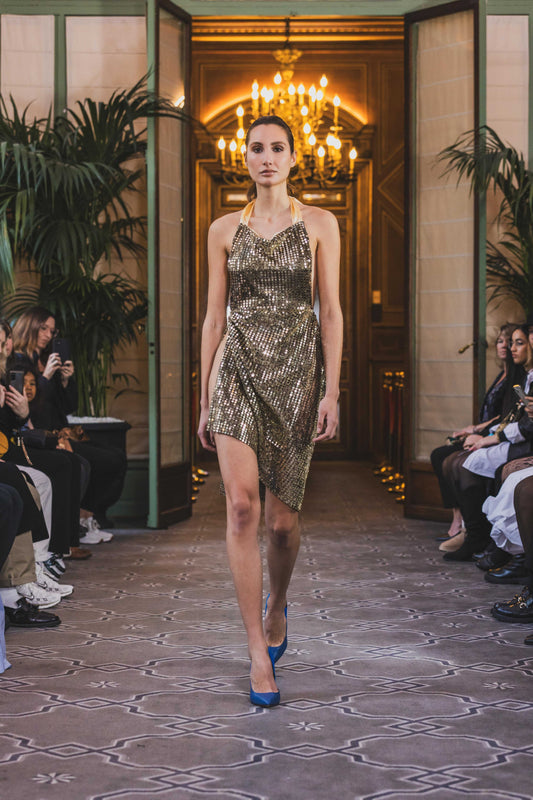 This photo shows a model wearing a size adjustable golden sequin dress with an asymmetrical skirt, a high thigh slit, a tie behind the neck, and satin lined bodice.