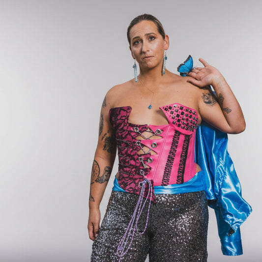 A model wearing a high-quality pink faux leather corset with black lace, studs, and rhinestones, paired with sequin pants and a blue fabric draped over one shoulder.
