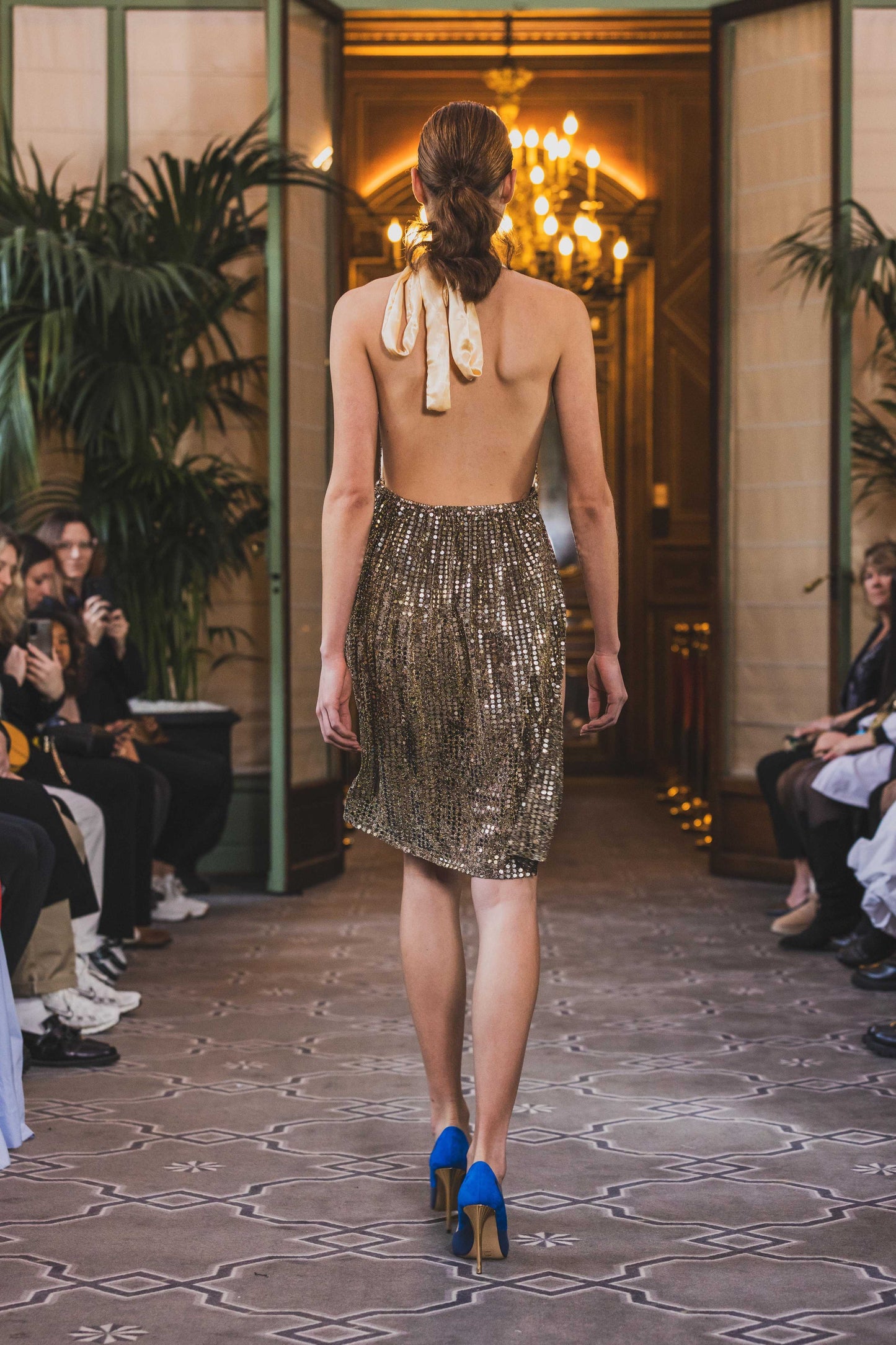 This photo shows a model wearing a size adjustable golden sequin dress with an asymmetrical skirt, a high thigh slit, a tie behind the neck, and satin lined bodice. 